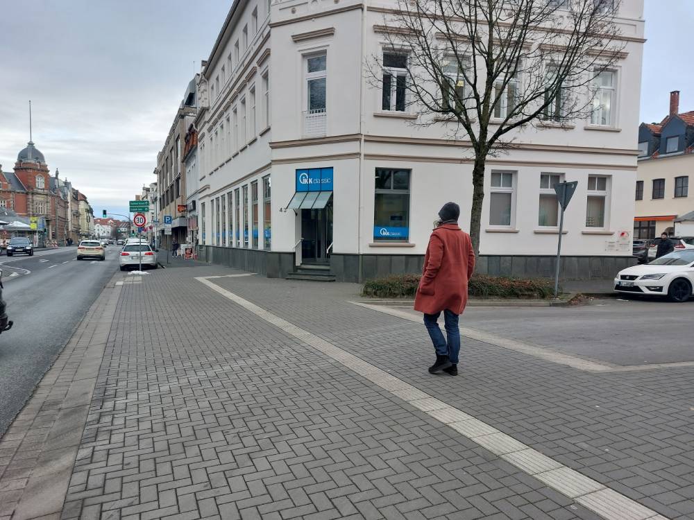 Gepflasterte Gehwegüberfahrt an einer Straßeneinmündung mit taktilem Leitsystem und Bordsteinkante, Fußgänger in rotem Mantel quert die Straße.