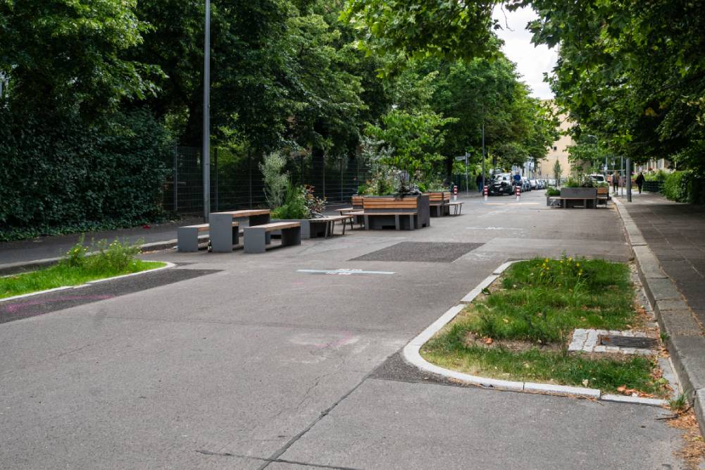 Straße mit mehreren braunen Holzbänken und Tischen auf der Fahrbahn, umgeben von Bäumen und Grünflächen, Berlin-Mitte.