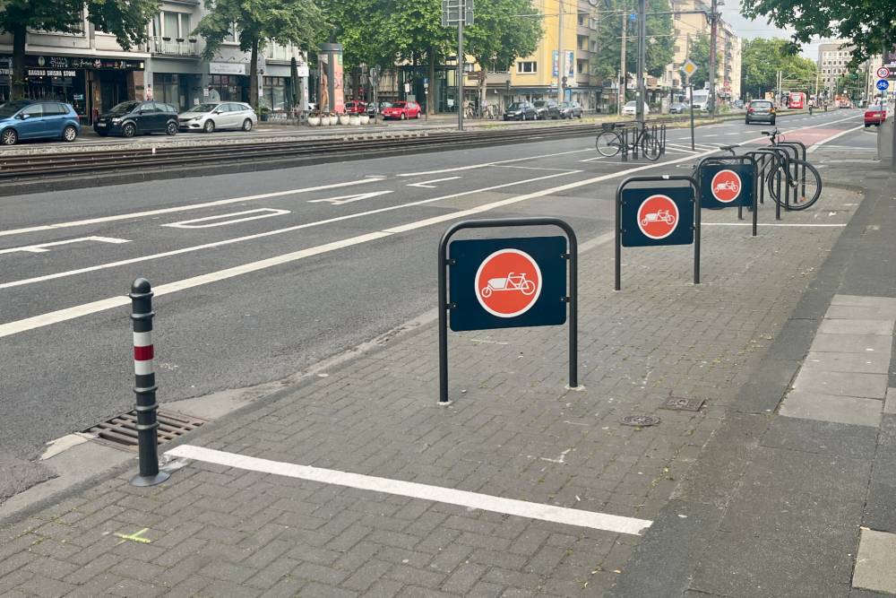 Mehrere Fahrradabstellplätze entlang eines Bürgersteigs an einer Straße. Die Abstellplätze sind durch schwarze Metallbügel mit einem orangefarbenen Schild gekennzeichnet, das ein weißes Lastenrad-Symbol zeigt. Im Hintergrund sind weitere Fahrradständer und einige geparkte Fahrräder zu sehen. Die Straße hat mehrere Fahrspuren und ist von Bäumen und Gebäuden gesäumt.