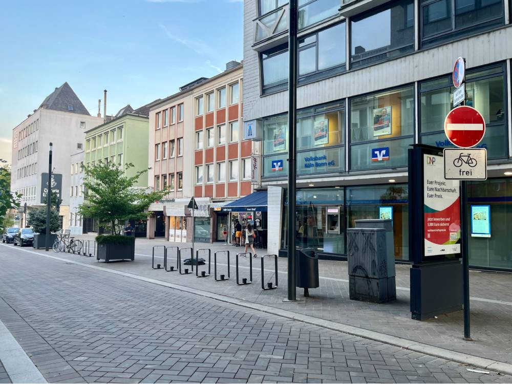 Eine städtische Straße in Köln mit einem Multifunktionsstreifen, der als Ladebereich für Kraftfahrzeuge, technische Infrastruktur, mobile Begrünung und Radabstellanlagen dient. Rechts im Bild steht ein Verkehrsschild mit einem roten Durchfahrtsverbotsschild und einem Zusatzschild mit einem Fahrradsymbol und dem Wort 'frei'. Entlang des Gehwegs befinden sich mehrere Fahrradständer und ein großer Pflanzkübel mit grünen Pflanzen. Im Hintergrund sind mehrstöckige Gebäude mit großen Fenstern und verschiedenen Fassadenfarben zu sehen, darunter ein modernes Glasgebäude und ältere, pastellfarbene Häuser. Die Straße ist mit grauen Pflastersteinen belegt, und es sind keine Fahrzeuge auf der Fahrbahn zu sehen.