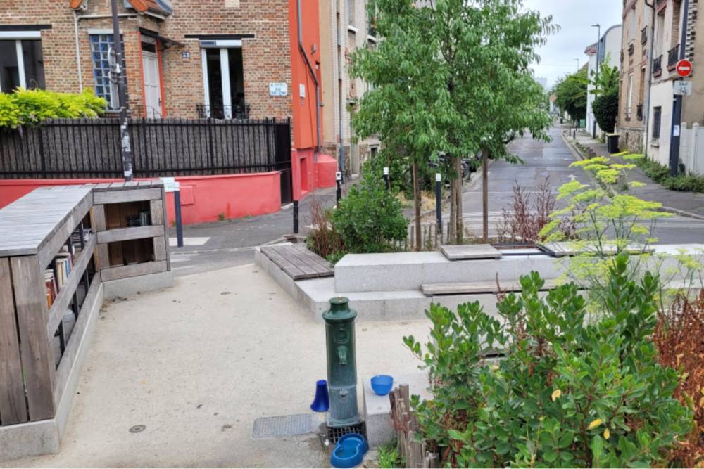 Ein "Pocket-Park" an der Rue Saigne in Montreuil zeigt einen ehemaligen Knotenpunkt, der entsiegelt und bepflanzt wurde. Im Vordergrund steht ein grüner Trinkbrunnen mit zwei blauen Bechern. Rechts davon befinden sich verschiedene Pflanzen und ein Baum, der in einem rechteckigen, niedrigen Betonbeet steht, das gleichzeitig als Sitzgelegenheit dient. Links ist eine Holzbank mit Rückenlehne zu sehen, die an eine niedrige Holzwand grenzt. Im Hintergrund verläuft eine Straße mit weiteren Gebäuden und Bäumen. Der Bereich wirkt als linearer Modalfilter und bietet Möglichkeiten zum Verweilen.