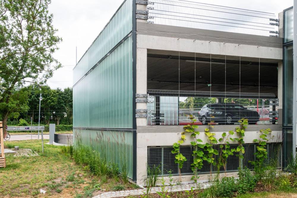 Mehrstöckige Quartiersgarage mit sichtbaren Parkplätzen, umgeben von vertikalen Begrünungselementen an der Fassade. Die Garage besteht aus Beton und hat teilweise transparente, grünlich getönte Fassadenelemente. Im Vordergrund wachsen verschiedene Pflanzen und Gras auf dem Boden. Im Hintergrund sind Bäume und ein Gehweg zu sehen.