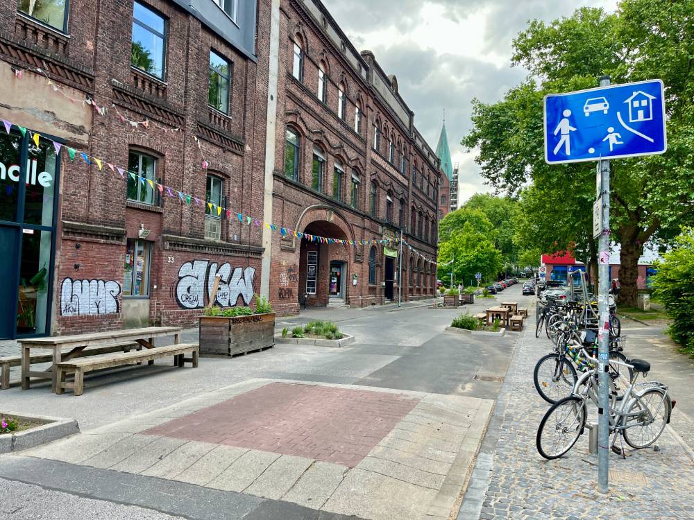 Eine ruhige Straße in Bochum mit einem mehrstöckigen Backsteingebäude auf der linken Seite, das mit Graffiti verziert ist. Vor dem Gebäude stehen Holzbänke und Tische. Rechts im Bild sind mehrere Fahrräder an einem Fahrradständer geparkt. Ein blaues Verkehrsschild zeigt Symbole für Fußgänger, spielende Kinder, ein Auto und ein Haus, was auf einen verkehrsberuhigten Bereich hinweist. Im Hintergrund sind grüne Bäume und ein Kirchturm sichtbar. Der Himmel ist bewölkt.