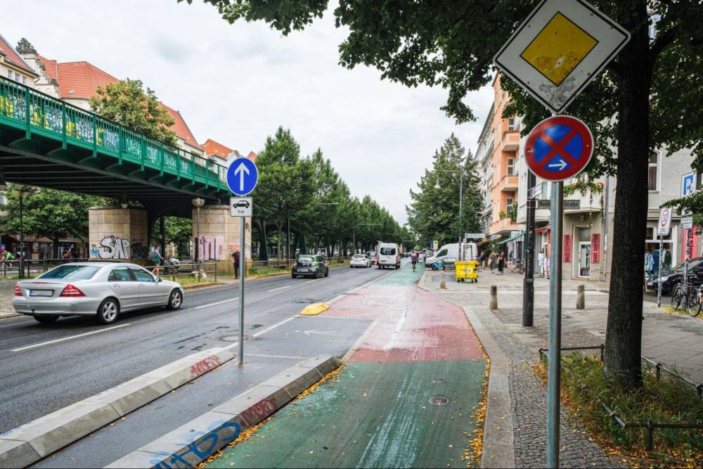 Die Aufnahme zeigt einen Straßenabschnitt der Schönhauser Allee in Berlin mit einem geschützten Radfahrstreifen, der durch eine bauliche Trennung vom Autoverkehr abgegrenzt ist. Direkt neben dem Radfahrstreifen befindet sich ein Wartebereich für Fußgänger, der durch eine grün und rot markierte Fläche auf dem Boden erkennbar ist. Rechts am Straßenrand stehen mehrere Verkehrsschilder, darunter ein blaues Schild mit weißem Pfeil nach oben, ein gelbes Dreieck mit weißem Rand und ein rundes Schild mit rotem Rand und blauem Hintergrund, das ein Halteverbot anzeigt. Links im Bild verläuft eine Hochbahnstrecke auf einer grünen Stahlbrücke über die Straße. Im Hintergrund sind Bäume und mehrstöckige Wohngebäude zu sehen.