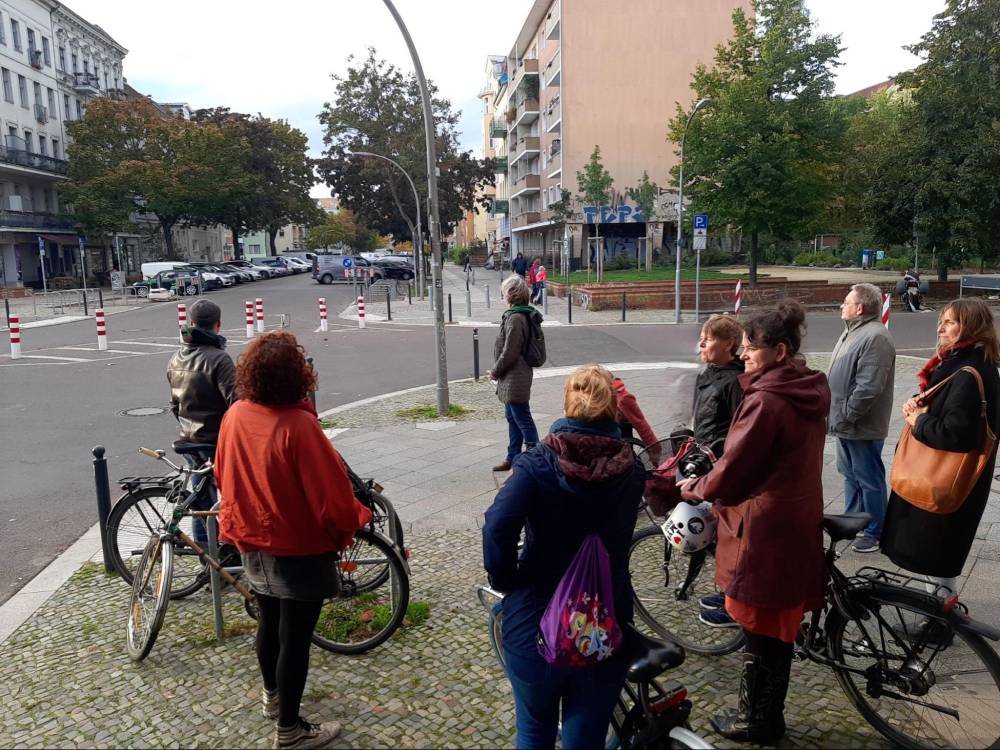 Mehrere Personen stehen mit Fahrrädern an einer Straßenecke in einem Wohngebiet mit mehrstöckigen Gebäuden und Bäumen.