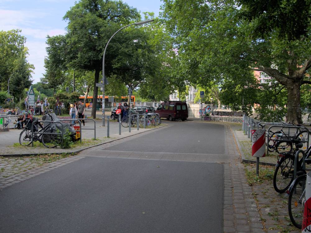 Eine Straße mit einer Aufpflasterung und Fahrbahneinengung, die zur Temporeduktion dient. Rechts und links der Straße sind Fahrradständer mit mehreren abgestellten Fahrrädern zu sehen. Im Hintergrund stehen Menschen, einige davon auf dem Gehweg, der von Bäumen gesäumt ist. Am rechten Straßenrand befindet sich ein rot-weißes Warnschild. Die Straße ist von Bäumen umgeben, die Schatten auf die Fahrbahn werfen. Im Hintergrund ist ein orangefarbener Bus zu erkennen. Die Szene spielt in der Weichselstraße in Berlin Friedrichshain-Kreuzberg vor einem Schuleingang mit starkem Fußgängerquerungsbedarf.