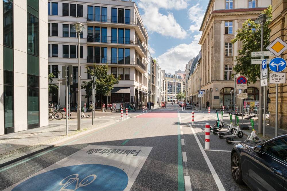 Die Fahrradstraße Oberwallstraße in Berlin ist eine breite Straße mit vorgezogenen Seitenräumen, die durch weiße Poller von der Fahrbahn getrennt sind. Die Fahrbahn ist in der Mitte rot markiert, was den Bereich für Fahrräder hervorhebt. Auf der rechten Straßenseite befinden sich mehrere E-Scooter, die auf einem definierten Bereich abgestellt sind. Links sind mehrere Fahrräder an einem Fahrradständer geparkt. Die Straße ist von mehrstöckigen Gebäuden mit großen Fenstern gesäumt. Verkehrsschilder weisen auf die Fahrradstraße und auf die erlaubte Nutzung durch Fahrräder und Anlieger hin. Der Himmel ist teilweise bewölkt.