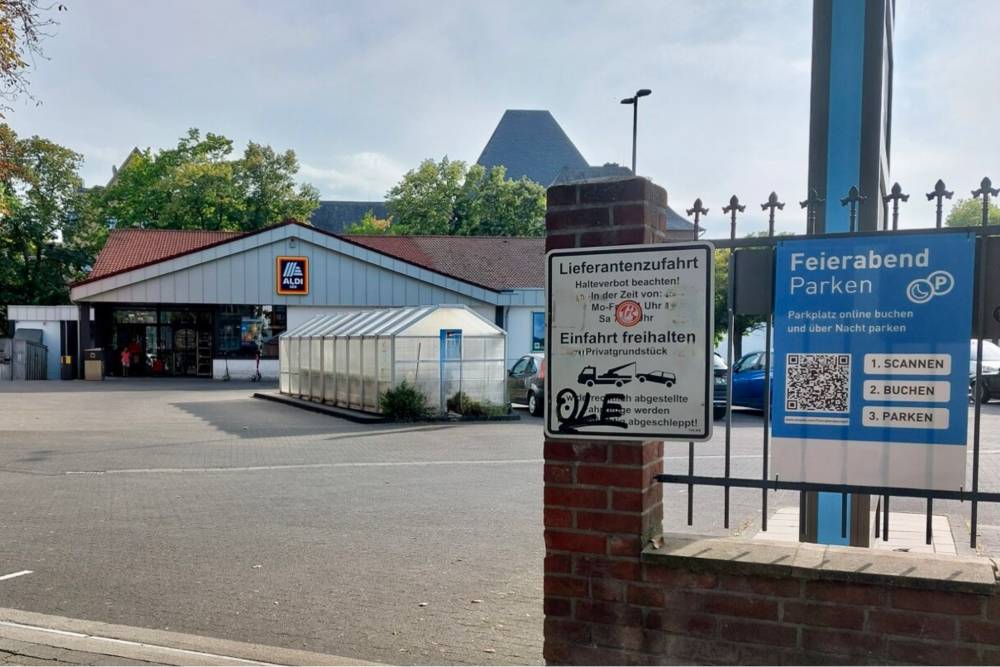 Vor einem Gewerbegebäude mit rotem Satteldach und weißer Fassade steht ein großer, weißer Container auf einem Parkplatz. Im Vordergrund ist ein schwarzes Metalltor mit zwei Schildern befestigt: Das linke Schild weist auf eine Lieferantenzufahrt hin und warnt, die Einfahrt freizuhalten, da sonst abgeschleppt wird. Das rechte Schild informiert über das Feierabend-Parken mit den Schritten '1. SCANNEN', '2. BUCHEN' und '3. PARKEN' sowie einem QR-Code. Im Hintergrund sind Bäume und ein wolkenverhangener Himmel zu sehen.