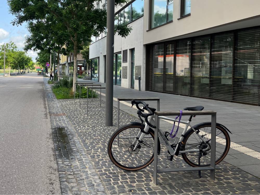 Ein silbernes Fahrrad ist an einem rechteckigen, aus Metall gefertigten Fahrradständer auf einem gepflasterten Gehweg angeschlossen. Der Gehweg besteht aus kleinen, quadratischen Pflastersteinen, die in einem regelmäßigen Muster verlegt sind. Rechts neben dem Fahrradständer verläuft ein breiter, glatter Gehweg mit dunklen Bodenplatten. Im Hintergrund sind moderne Gebäude mit großen Fenstern und Rollläden zu sehen. Links neben dem Gehweg verläuft eine Straße, die von Bäumen gesäumt ist. Die Straße ist frei von parkenden Kraftfahrzeugen. Der Himmel ist teilweise bewölkt, und die Szene wirkt ruhig und aufgeräumt.