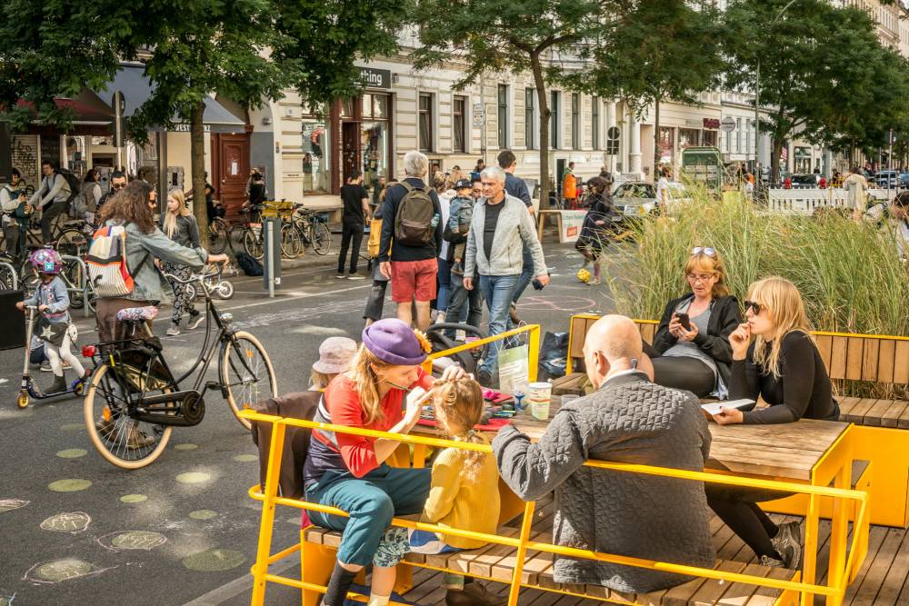 In der Bergmannstraße befindet sich ein Parklet mit gelben Sitzgelegenheiten und einem Holztisch, umgeben von bepflanzten Bereichen mit hohem Gras. Menschen sitzen entspannt auf den Bänken, einige unterhalten sich, andere beschäftigen sich mit Kindern. Im Hintergrund sind weitere Personen zu sehen, die zu Fuß oder mit dem Fahrrad unterwegs sind. Die Straße ist verkehrsberuhigt gestaltet, mit Bäumen entlang des Gehwegs und Wohngebäuden im Hintergrund. Auf dem Asphalt sind bunte Kreidezeichnungen sichtbar. Die Szene vermittelt eine lebendige Begegnungszone.