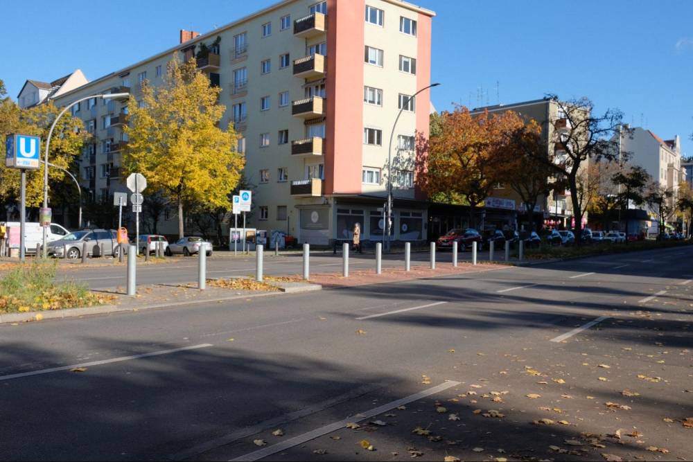 Eine sichere Querungsstelle für Fuß- und Radverkehr im Mittelstreifen einer Hauptverkehrsstraße. Der Mittelstreifen ist durch mehrere weiße Poller abgegrenzt, die das Überqueren erleichtern und den Verkehr trennen. Im Hintergrund sind mehrstöckige Wohngebäude mit herbstlich gefärbten Bäumen zu sehen. Am linken Bildrand steht ein blaues U-Bahn-Schild, das auf eine nahegelegene U-Bahn-Station hinweist. Die Straße ist breit und asphaltiert, auf dem Boden sind keine sichtbaren Markierungen für die Querung zu erkennen.