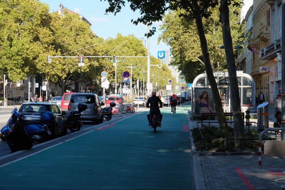 Ein grün markierter, baulich getrennter Zweirichtungs-Radweg entlang der Praterstraße in Wien, der durch einen Parkstreifen zusätzlich vom Autoverkehr geschützt ist. Auf dem Radweg fahren zwei Radfahrer in einigem Abstand voneinander. Rechts neben dem Radweg verläuft ein Gehweg mit Bäumen und einer Bushaltestelle mit einem Werbeplakat. Links neben dem Radweg sind parkende Autos und Motorräder zu sehen. Im Hintergrund sind Ampeln und ein U-Bahn-Symbol sichtbar. Die Straße ist von Bäumen gesäumt, die Schatten auf den Radweg werfen.