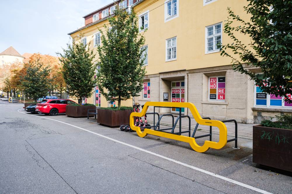Eine gelb lackierte Fahrradabstellanlage in Form eines Autos steht auf einem ehemaligen Kfz-Stellplatz an einem Bürgersteig. Die Anlage besteht aus einem Metallrahmen, der die Umrisse eines Autos nachzeichnet, und mehreren Fahrradständern im Inneren. Im Hintergrund sind ein gelb gestrichenes Wohngebäude mit mehreren Fenstern und Bäumen zu sehen. Auf dem Bürgersteig stehen zudem große Pflanzkübel mit Bäumen. Ein rotes Auto ist auf einem angrenzenden Parkplatz geparkt.