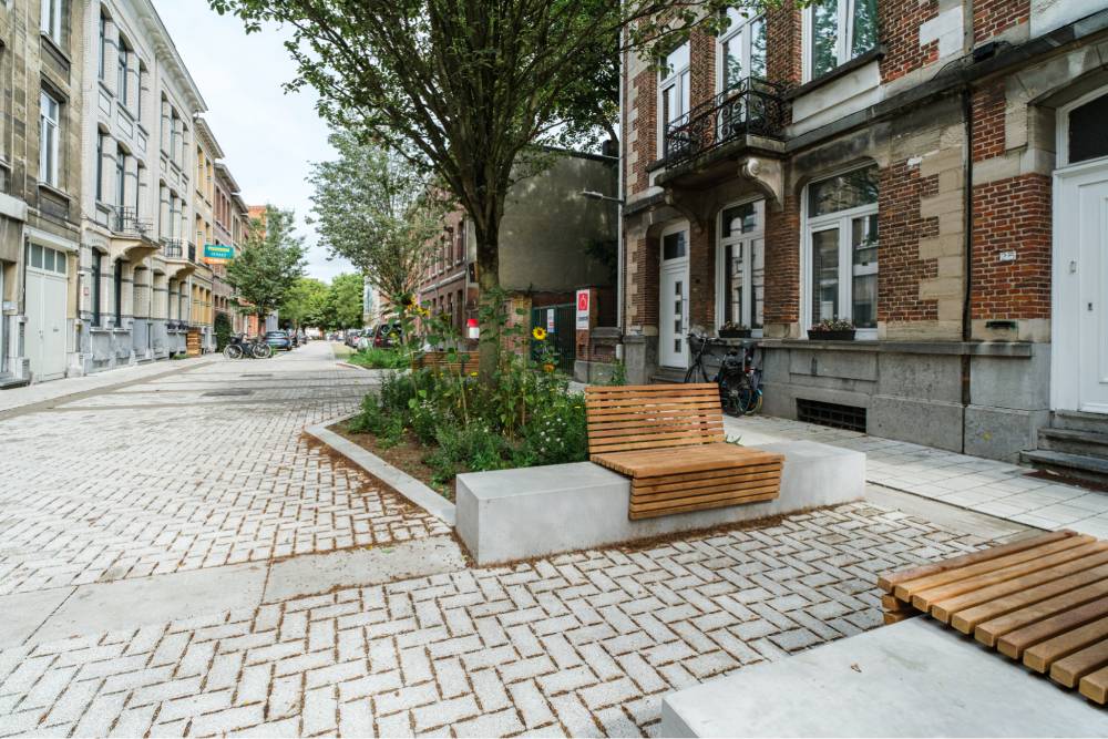 Der Eingangsbereich eines Mehrfamilienhauses in Antwerpen zeigt einen halböffentlichen Raum mit vorgelagerten Grünflächen und Sitzgelegenheiten. Im Vordergrund steht eine Sitzbank aus Holz mit einer Rückenlehne, die auf einem rechteckigen, hellgrauen Betonsockel montiert ist. Daneben befindet sich eine weitere Sitzgelegenheit aus Holz ohne Rückenlehne. Die Fläche ist mit hellen, rechteckigen Pflastersteinen in einem Fischgrätmuster belegt. Im Hintergrund sind Bäume und weitere Grünpflanzen zu sehen, die den Bereich begrenzen. Die angrenzenden Gebäude sind aus Backstein mit weißen Fensterrahmen und Türen. Die Straße verläuft links neben dem Grünbereich und ist ebenfalls gepflastert.