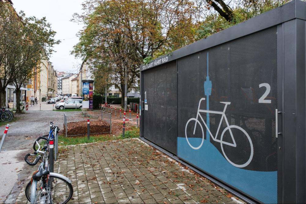 Eine Sammelschließanlage für Fahrräder steht auf einem gepflasterten Platz in einem Wohnquartier in Stuttgart. Die Anlage ist rechteckig, schwarz und hat eine perforierte Metallfront, auf der ein großes weißes Fahrradsymbol und die Zahl '2' zu sehen sind. Darüber steht in weißen Buchstaben 'FAHRT RAD'. Im Vordergrund sind zwei Fahrräder an einem Fahrradständer angeschlossen. Im Hintergrund sind mehrstöckige Wohngebäude, Bäume mit herbstlichem Laub und ein geparkter weißer Pkw zu sehen. Der Himmel ist bewölkt.
