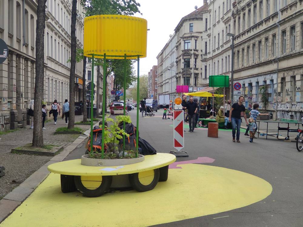 In einer Straße im ersten Kiezblock in Leipzig stehen moderne Sitzgelegenheiten und Stadtmöbel. Im Vordergrund befindet sich eine runde, gelb lackierte Sitzbank mit einer integrierten bepflanzten Fläche in der Mitte. Über der Bank ist ein gelber, zylindrischer Sonnenschutz angebracht, der von grünen Stangen getragen wird. Im Hintergrund sind weitere Sitzgelegenheiten mit ähnlichen Sonnenschutzstrukturen in verschiedenen Farben wie Pink und Grün zu sehen. Die Straße ist von mehrstöckigen Altbauten gesäumt, und mehrere Personen gehen oder sitzen auf den Sitzmöbeln. Der Boden um die Sitzbank ist ebenfalls gelb markiert, wodurch der Bereich optisch hervorgehoben wird.