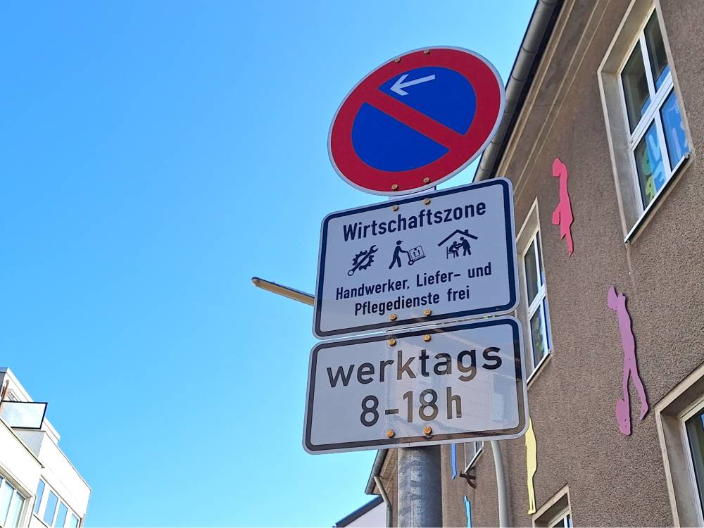 Ein Verkehrsschild mit einem roten Kreis und einem blauen Hintergrund, durch den ein roter Querbalken verläuft, zeigt ein Parkverbot mit Pfeil nach links an. Darunter befinden sich zwei weiße Zusatzschilder mit schwarzer Schrift. Das obere Zusatzschild trägt die Überschrift 'Wirtschaftszone' und zeigt drei Piktogramme: einen Hammer und Schraubenschlüssel, ein Lieferfahrzeug und eine Person, die eine andere Person pflegt. Darunter steht der Text 'Handwerker, Liefer- und Pflegedienste frei'. Das untere Zusatzschild gibt die zeitliche Beschränkung 'werktags 8-18 h' an. Im Hintergrund ist eine Hausfassade mit mehreren Fenstern und pinkfarbenen, abstrakten Figuren an der Wand zu sehen. Der Himmel ist klar blau.