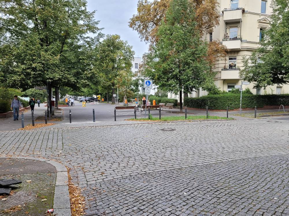Blick auf eine zurückgebaute Einmündung mit Kopfsteinpflaster, Bäumen, mehreren Pollern und einem Fußgänger mit Fahrrad im Berliner Komponistenviertel.