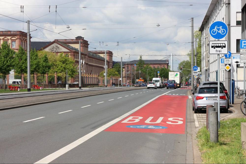 Die Bismarckstraße in Mannheim zeigt eine Umweltspur, die durch eine rote Fahrbahnmarkierung mit dem weißen Schriftzug 'BUS' gekennzeichnet ist. Die Umweltspur befindet sich am rechten Fahrbahnrand und ist für Busse und Radverkehr reserviert. Ein blaues Verkehrsschild mit einem weißen Fahrradsymbol und dem Zusatz 'frei' erlaubt Radfahrern die Nutzung der Spur. Rechts neben der Umweltspur sind parkende Autos zu sehen. Links und rechts der Straße stehen mehrstöckige Gebäude, links sind sie von Bäumen gesäumt.