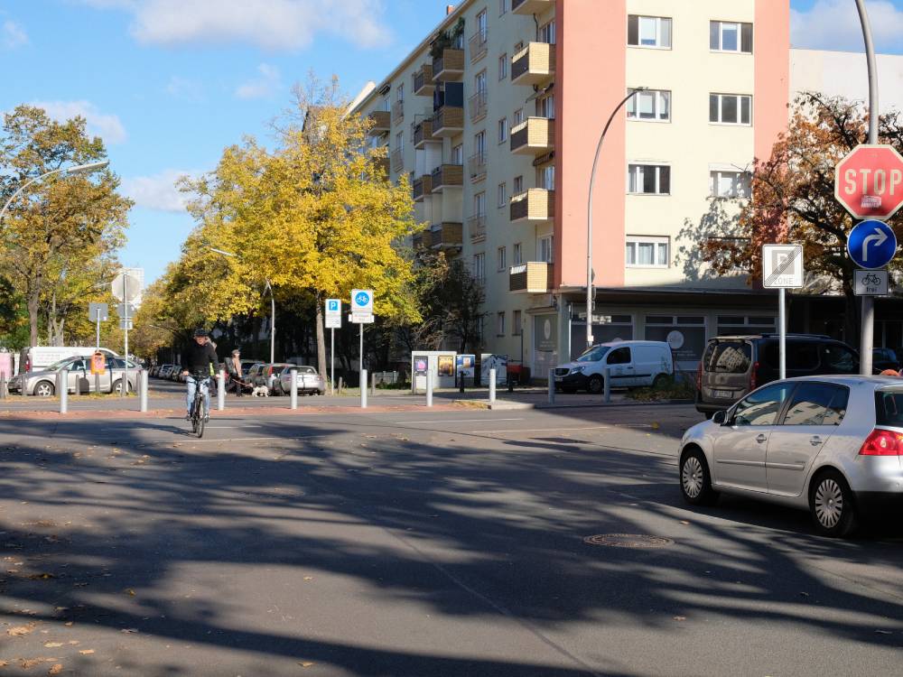 Kreuzung in Berlin mit Fahrradstraße, Radfahrer auf der Straße, rechts abbiegende Autos, Stoppschild und Abbiegepfeil auf blauem Schild.