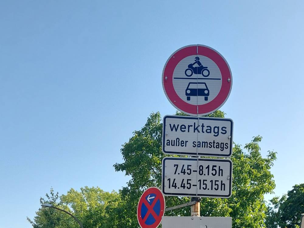 Ein rundes Verkehrszeichen mit rotem Rand zeigt ein Motorrad über einem Auto, was auf ein Fahrverbot für diese Fahrzeugarten hinweist. Darunter befinden sich drei rechteckige Zusatzschilder mit schwarzer Schrift auf weißem Grund. Das erste Zusatzschild trägt die Aufschrift 'werktags außer samstags', das zweite zeigt die Zeiten '7.45 - 8.15 h' und '14.45 - 15.15 h'. Im Hintergrund sind grüne Baumkronen und ein blauer Himmel zu sehen.