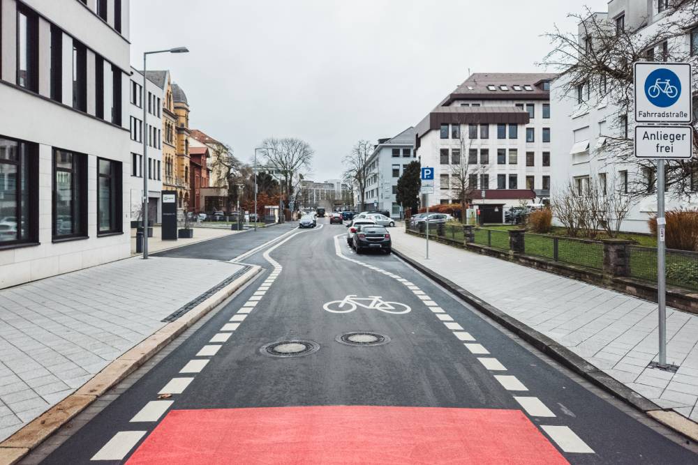 Eine Fahrradstraße in Kassel mit einem roten Sicherheitstrennstreifen auf der Fahrbahn, der von weißen, gestrichelten Linien eingerahmt ist. Auf dem Asphalt ist ein weißes Fahrradsymbol aufgemalt. Rechts am Straßenrand steht ein blau-weißes Verkehrsschild mit dem Symbol eines Fahrrads und der Aufschrift 'Fahrradstraße' sowie einem Zusatzschild mit der Aufschrift 'Anlieger frei'. Links und rechts der Straße sind moderne Wohn- und Geschäftshäuser zu sehen, auf der rechten Seite sind parkende Autos entlang der Straße abgestellt.