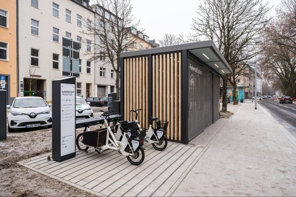 Eine moderne Mobilitätsstation in Düsseldorf mit einer Sammelschließanlage für Fahrräder, erkennbar an einem länglichen, rechteckigen Bau mit vertikalen Holzlatten an der Seite und einer grauen Front. Vor der Anlage stehen zwei weiße Lastenräder mit schwarzem Gepäckträger und Aufbauten. Links neben den Lastenrädern befindet sich ein weißer Informations- oder Bedienautomat mit der Aufschrift 'Lastenrad Automat'. Im Hintergrund sind mehrstöckige Wohngebäude und eine Straße mit parkenden Autos zu sehen.