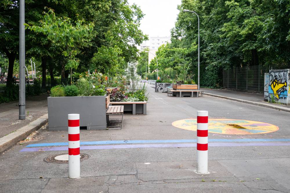Eine breite, asphaltierte Straße in Berlin-Mitte ist durch mehrere große, graue Pflanztröge mit grünen Pflanzen und kleinen Büschen in der Mitte geteilt. Am Straßenrand stehen Bänke aus Holz mit Metallgestellen. Im Vordergrund sind zwei weiße Poller mit roten Streifen auf der Straße zu sehen, die den Zugang für Fahrzeuge verhindern. Rechts auf der Straße ist ein bunter, kreisförmiger Bodenaufdruck mit gelben, roten und blauen Elementen. Links und rechts der Straße stehen hohe, grüne Bäume, die den Bereich beschatten. Im Hintergrund sind mehrstöckige Wohngebäude sichtbar.