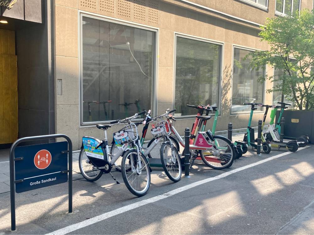 Vor einem Gebäude mit großen Fenstern stehen mehrere Leihräder und E-Scooter nebeneinander auf einem Bürgersteig. Die Fahrräder sind mit verschiedenen Werbeaufklebern versehen, darunter ein rotes Fahrrad mit dem Logo der Telekom. Rechts davon befinden sich mehrere grün-schwarze E-Scooter. Links steht ein blaues Schild mit einem orangefarbenen Symbol und der Aufschrift 'Große Sandkaul'. Die Szene zeigt eine Sharingstation in der Kölner Altstadt.