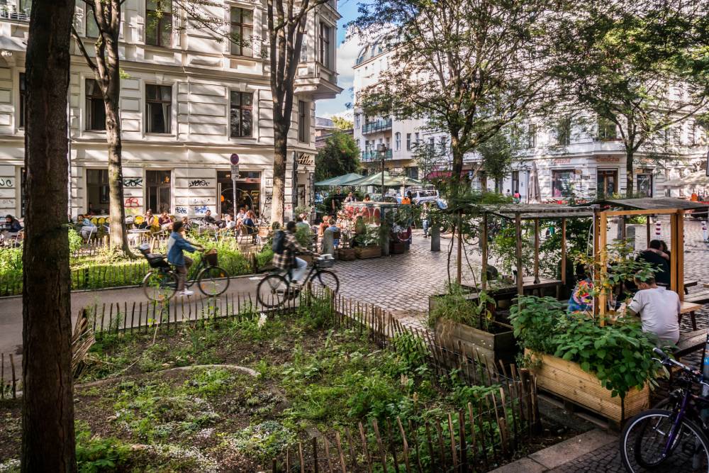 In einem urbanen Garten im Graefekiez in Berlin sind verschiedene Beete mit Pflanzen und Gemüse zu sehen, die von kleinen Holzzäunen umgeben sind. Im Vordergrund steht ein rechteckiges Hochbeet mit grünen Pflanzen, daneben sitzt eine Person an einem Tisch unter einem kleinen Pavillon. Im Hintergrund sind mehrere Menschen auf Fahrrädern unterwegs, umgeben von Bäumen und mehrstöckigen Altbauten mit weißen Fassaden und zahlreichen Fenstern. Die Szene zeigt eine belebte, grüne Stadtecke mit urbaner Begrünung und sozialer Aktivität.