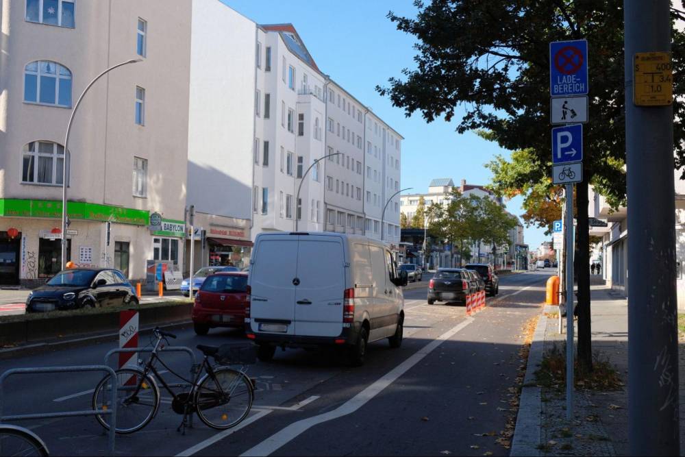 Die Aufnahme zeigt die Grunewaldstraße in Berlin-Schöneberg mit einer kombinierten Lade- und Lieferzone auf der rechten Straßenseite. Dort steht ein weißer Lieferwagen in der Ladezone, die durch weiße Markierungen auf dem Asphalt gekennzeichnet ist. Vor der Ladezone befindet sich eine Fußgänger-Aufstellfläche, die durch eine weiße Linie vom Radfahrstreifen getrennt ist. Der Radfahrstreifen ist durch eine durchgezogene weiße Linie und Fahrradsymbole auf dem Asphalt sichtbar. Am rechten Straßenrand stehen mehrere Verkehrsschilder, darunter ein blaues Schild mit dem Text 'Ladebereich' und ein Parkzeichen mit einem Fahrradsymbol. Im Vordergrund links sind zwei Fahrräder an Anlehnbügeln angeschlossen. Die Straße ist von mehrstöckigen Wohngebäuden gesäumt, und es sind einige Bäume mit grünem Laub zu sehen. Die Szene wirkt sonnig und klar, mit Schatten auf dem Asphalt.