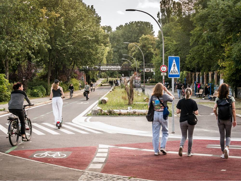 Eine Fahrradstraße mit gesondertem Fußgängerbereich am Görlitzer Ufer im Wrangelkiez in Berlin. Im Vordergrund verläuft ein rot markierter Radweg mit einem weißen Fahrradsymbol. Rechts davon befindet sich ein breiter Fußweg, auf dem drei Personen in Richtung Straße gehen. Links fährt eine Person mit dem Fahrrad, eine weitere Person fährt auf einem E-Scooter. Im Hintergrund sind weitere Radfahrer und Fußgänger zu sehen. Die Straße ist von Bäumen gesäumt, und es gibt eine Fußgängerüberquerung mit einem blauen Verkehrsschild, das einen Zebrastreifen anzeigt. Straßenlaternen mit gebogenen Masten stehen entlang der Straße. Im Hintergrund führt eine Fußgängerbrücke über die Straße.