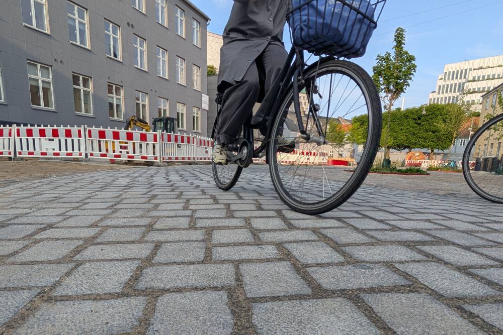 Das Bild zeigt eine Nahaufnahme eines geschliffenen Pflasters auf einem Platz in Aarhus, das aus rechteckigen, hellgrauen Steinen besteht, die in einem regelmäßigen Muster verlegt sind. Im Vordergrund ist ein Fahrrad mit einem schwarzen Rahmen und einem blauen Korb am Lenker zu sehen, dessen Vorderrad über das Pflaster rollt. Die Person auf dem Fahrrad trägt eine dunkelgraue Hose und schwarze Schuhe. Im Hintergrund sind mehrstöckige Gebäude mit hellen Fassaden sowie Absperrungen mit rot-weißen Streifen zu erkennen. Weiter hinten stehen grüne Bäume und ein modernes, weißes Gebäude mit vielen Fenstern.