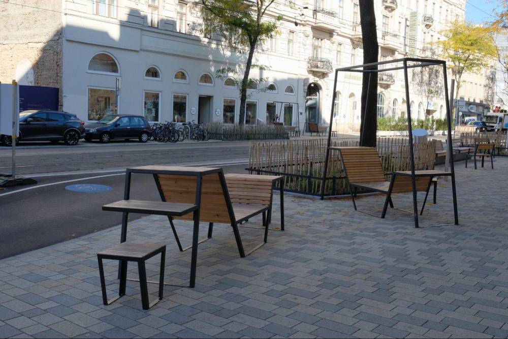 Entlang der Mariahilfer Straße in Wien sind moderne Sitzgelegenheiten aus Holz und schwarzem Metall auf einem gepflasterten Gehweg angeordnet. Die Sitzmöbel bestehen aus einer Kombination von Bänken mit Rückenlehnen, einer Bank mit Armlehnen und einem kleinen Hocker. Zwischen den Sitzgelegenheiten befinden sich Pflanzbeete mit jungen Bäumen, die von einem niedrigen Metallzaun umgeben sind. Im Hintergrund sind historische Gebäude mit weißen Fassaden und großen Fenstern zu sehen, sowie geparkte Autos und Fahrräder. Die Szene ist von Tageslicht erhellt, wobei Schatten auf den Gehweg fallen.