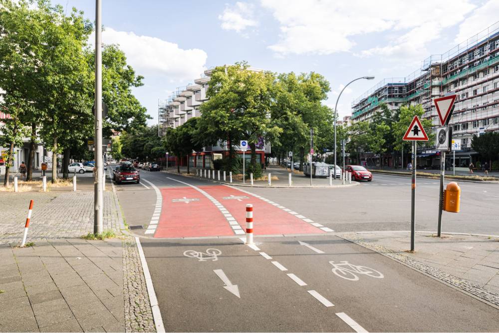 Eine Fahrradstraße in Berlin mit rotem, markiertem Radweg, der von einer selbständigen Führung in die Fahrradstraße übergeht. Der Radweg ist zweispurig mit weißen Fahrradsymbolen und Pfeilen auf dem Asphalt, die die Fahrtrichtung anzeigen. Am Übergang sind weiße Pfosten aufgestellt, die den Radweg vom restlichen Straßenbereich abgrenzen. Rechts neben dem Radweg steht ein Vorfahrts-Verkehrsschild. Im Hintergrund sind mehrstöckige Wohngebäude und Bäume zu sehen. Die Straße ist asphaltiert, der Gehweg gepflastert. Die Szene zeigt eine klare Trennung und Bevorrechtigung des Radverkehrs an der Kreuzung.