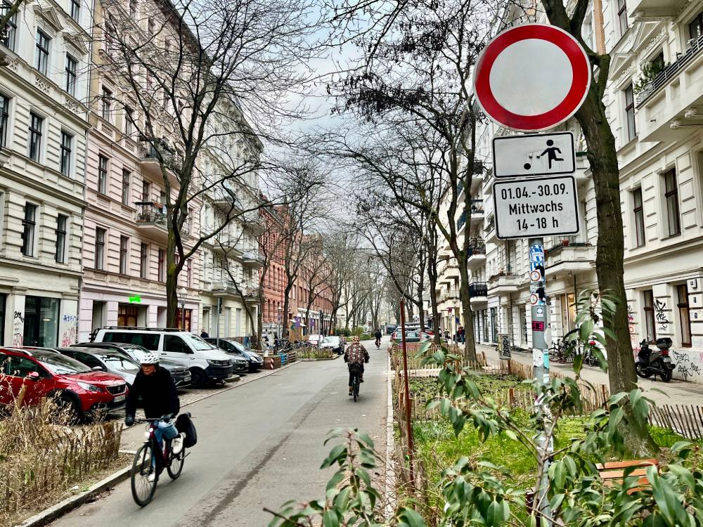 Eine temporäre Spielstraße im Berliner Graefekiez ist zu sehen. Die Straße ist von mehrstöckigen Altbauten mit weißen und roten Fassaden gesäumt. Rechts am Straßenrand steht ein Verkehrsschild mit einem roten Kreis, darunter ein Zusatzschild mit einem Piktogramm eines spielenden Kindes und der Aufschrift '01.04. - 30.09. Mittwochs 14 - 18 h'. Auf der Straße fahren zwei Personen mit dem Fahrrad in entgegengesetzte Richtungen. Links sind parkende Autos zu sehen, rechts gibt es einen schmalen Grünstreifen mit Bäumen und Büschen.