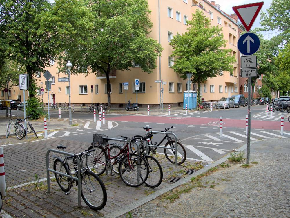 Vorgezogener Seitenraum an einer Kreuzung in der Jonasstraße/Ilsenstraße in Berlin-Neukölln, der durch weiße Markierungen auf der Fahrbahn und rot-weiße Pfosten abgegrenzt ist, um die Querungsdistanz für Fußgänger zu verringern. Im Vordergrund befinden sich mehrere Fahrräder, die an einem Fahrradständer angeschlossen sind. Rechts steht ein Verkehrsschild mit einem roten Dreieck (Vorfahrt gewähren) und einem blauen Pfeil nach oben, darunter zwei Zusatzschilder mit den Aufschriften 'Radverkehr frei' und 'Anlieger frei'. Im Hintergrund sind mehrstöckige Wohngebäude mit hellgelber Fassade und mehreren Fenstern zu sehen. Bäume mit grünem Laub säumen die Straße.