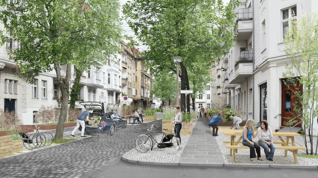 Ein ruhiger, begrünt gestalteter Straßenraum mit geglättetem Kopfsteinpflaster, das die Fahrbahnfläche bildet. Rechts befindet sich ein breiter Gehweg mit Sitzgelegenheiten in Form von Holzbänken und Tischen, auf denen zwei Frauen sitzen und sich unterhalten. Links sind Bäume gepflanzt, die Schatten spenden, und ein Fahrradständer mit mehreren Fahrrädern. Ein Auto steht mit geöffneter Heckklappe auf der Fahrbahn, daneben sind Menschen zu sehen, die sich bewegen oder Gegenstände tragen. Die Gebäude entlang der Straße sind mehrstöckig und haben helle Fassaden. Die Straße wirkt einladend und fördert Aufenthaltsqualität durch Begrünung und Sitzmöglichkeiten, während das Parken von Kraftfahrzeugen nur temporär erfolgt.